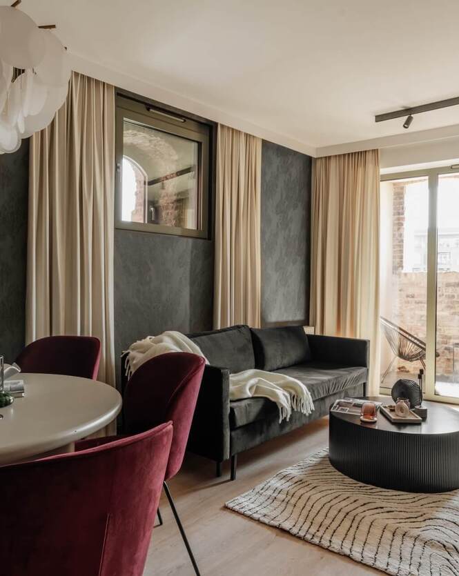Salonik apartamentu z szarą sofą, okrągłym stołem i bordowymi krzesłami, balkonem z dwoma fotelami i widokiem na ceglany mur.