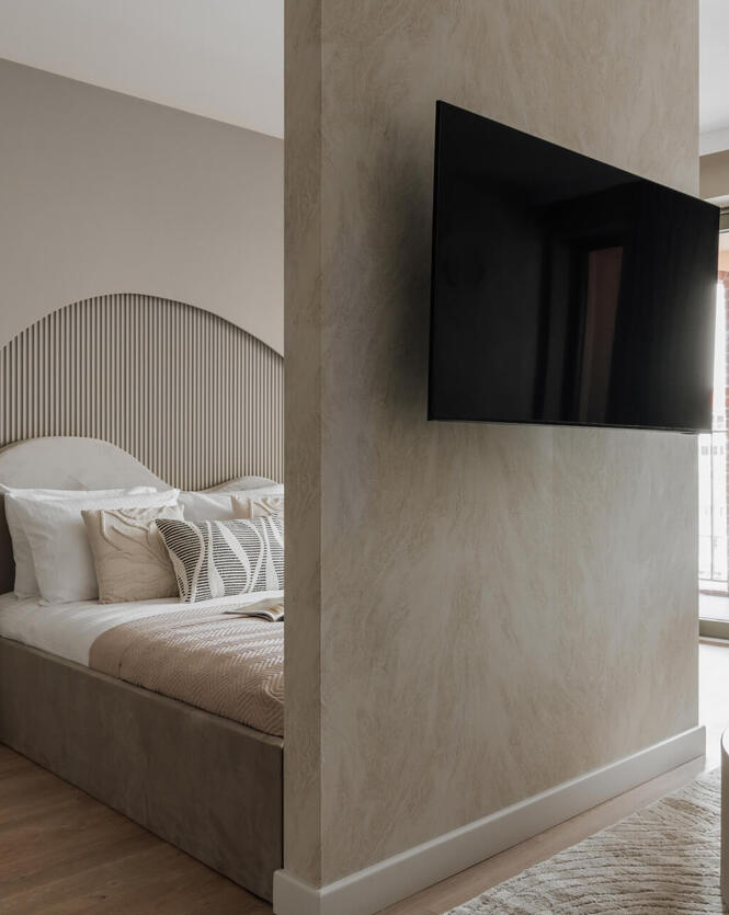 Przytulny pokój hotelowy z dużym łóżkiem, falistym tapicerowanym wezgłowiem, stolikiem nocnym z lampą, telewizorem ściennym i balkonem.