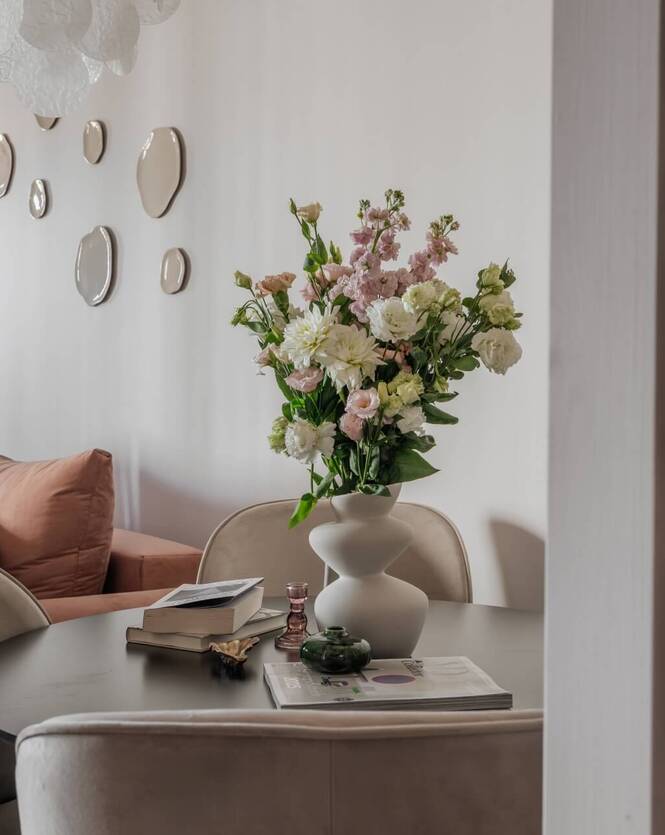 Przytulny salon z terakotową sofą, okrągłym stołem i wazą z pastelowymi kwiatami, ozdobne lustrzane plamy na ścianie.