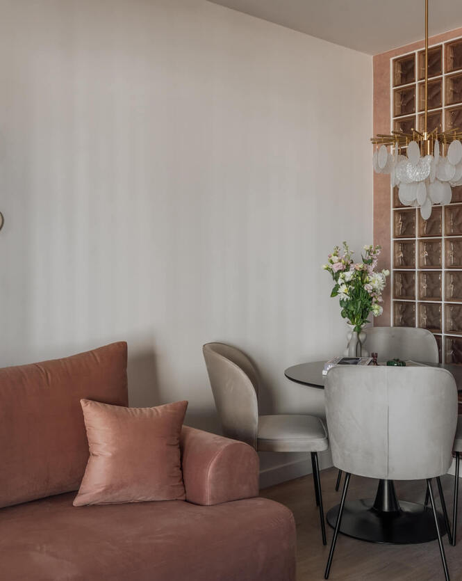 Kameralny lounge z terakotową sofą, okrągłym stolikiem i szarymi fotelami, szklaną ścianą dekoracyjną, ozdobną żyrandolową lampą i wazonem kwiatów.