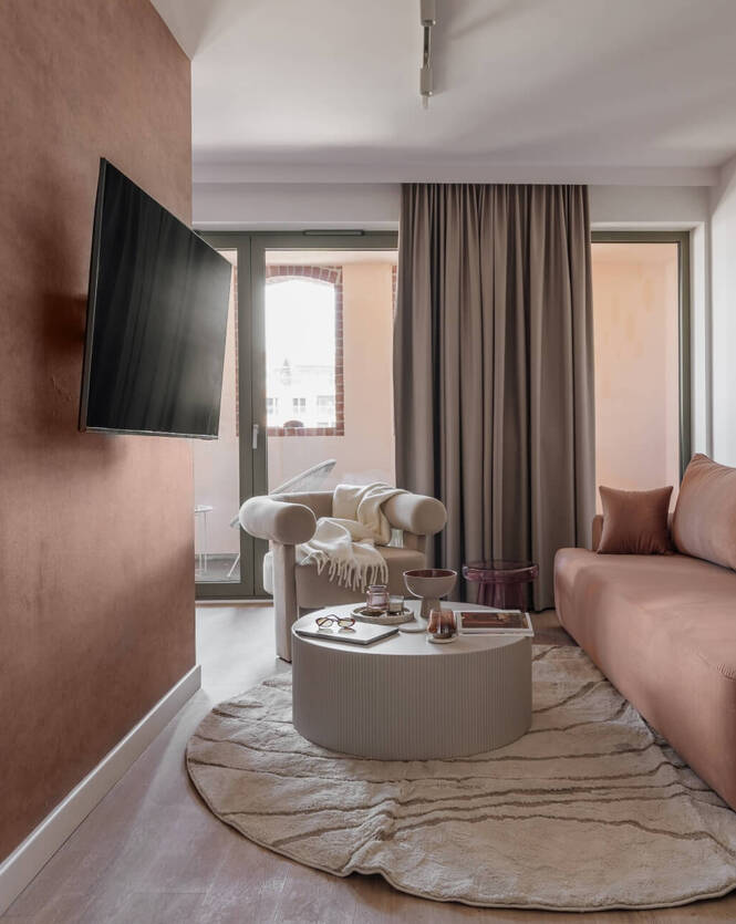 Przytulny apartament w ciepłych terakotowych tonach, sofa i fotel, oddzielone łóżko, balkon, TV na przegrodzie.