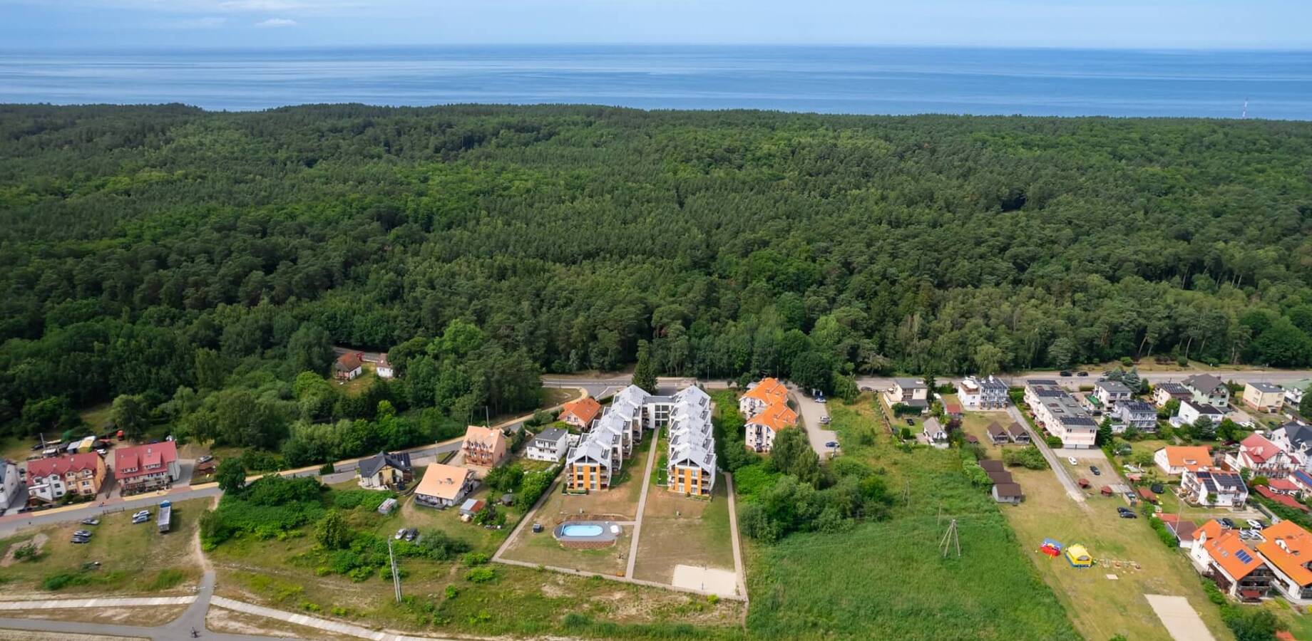Nadmorski kompleks apartamentów przy sosnowym lesie, z małym odkrytym basenem, promenadą i widokiem na morze.