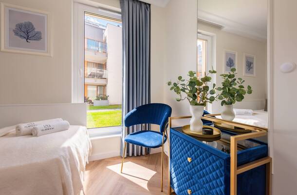 Jasny pokój hotelowy z łóżkiem i ręcznikami Flatbook, granatowym welurowym fotelem, złotą toaletką z lustrem, rośliną i widokiem na dziedziniec.