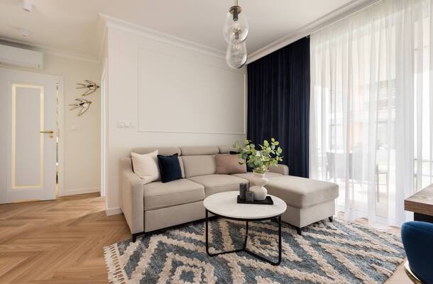 Przytulny salon apartamentu: beżowy narożnik z szezlongiem, marmurowy stolik z wazonem, zasłony, balkon i drewniana podłoga.