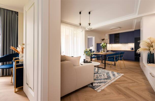 Nowoczesny apartament z otwartym salonem i jadalnią, granatową kuchnią, złotymi krzesłami, wiszącymi lampami i balkonem.