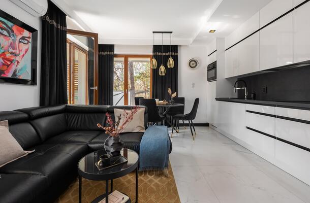 Nowoczesny apartament: biała kuchnia na wysoki połysk, czarna narożna sofa, złote lampy, jadalnia i balkon z drzewami.