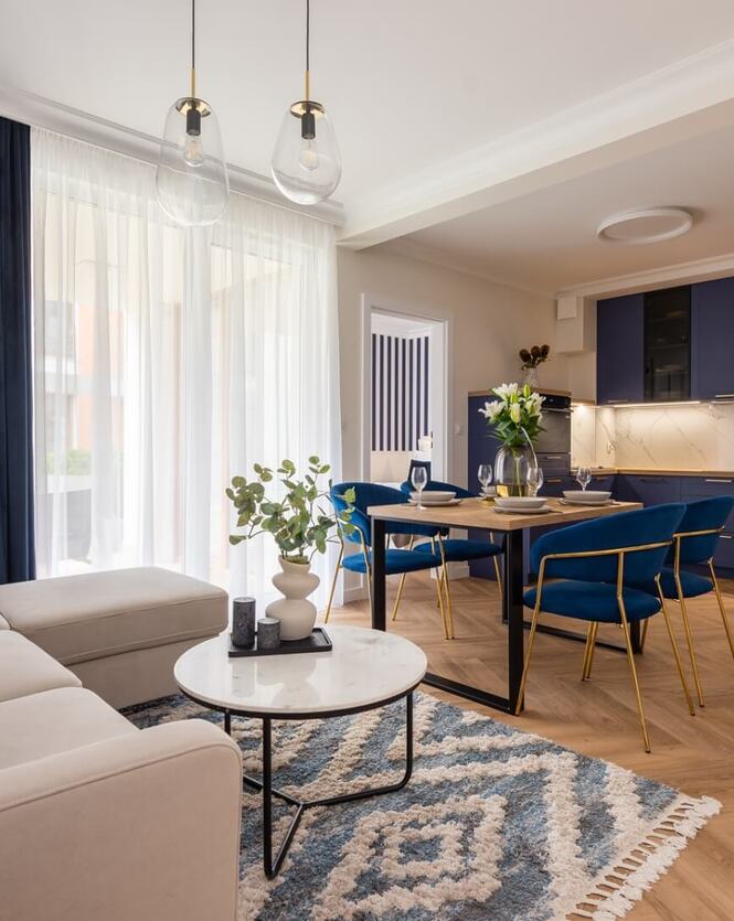 Elegancki apartament z aneksem kuchennym, jadalnią z granatowymi krzesłami, wygodną sofą, dużymi zasłonami i dekoracyjnymi lampami.