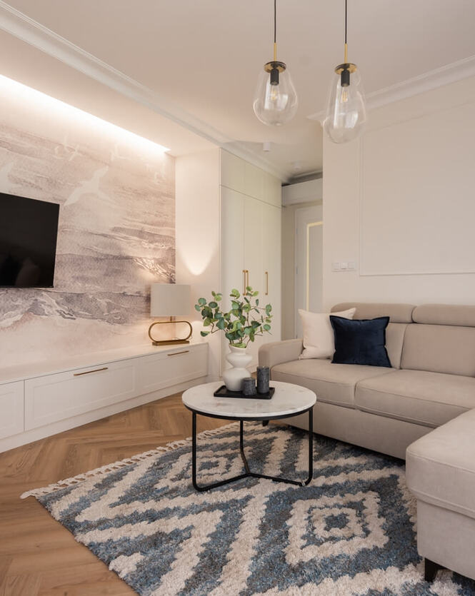 Przytulny salon apartamentu z beżowym narożnikiem, telewizorem na podświetlonej ścianie, stolikiem i dekoracjami.