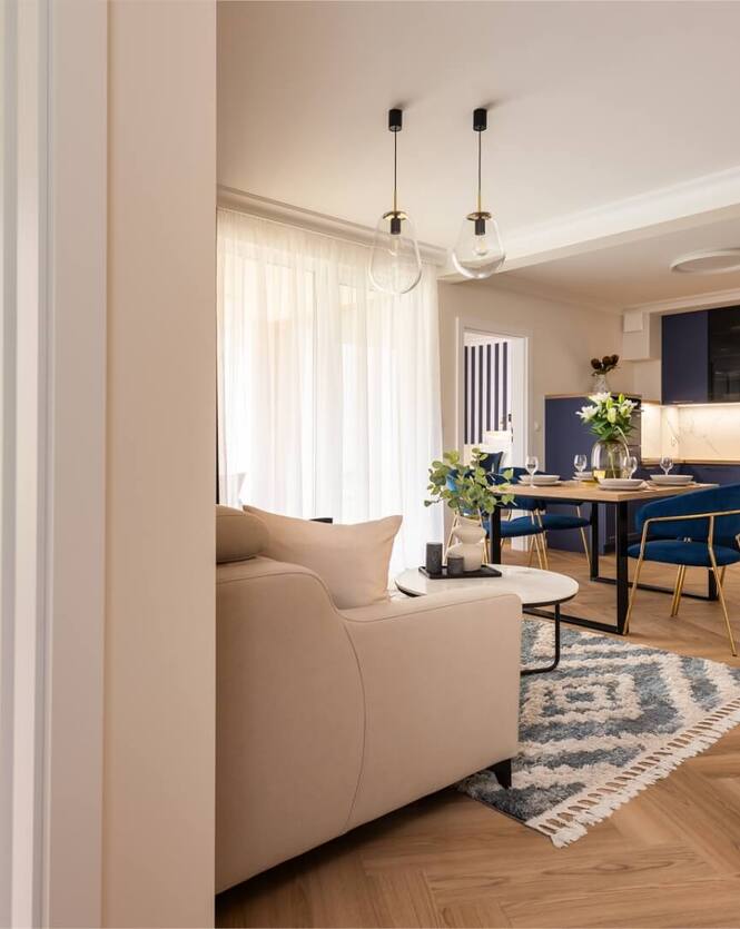 Nowoczesny apartament z otwartym salonem i jadalnią, granatową kuchnią, złotymi krzesłami, wiszącymi lampami i balkonem.