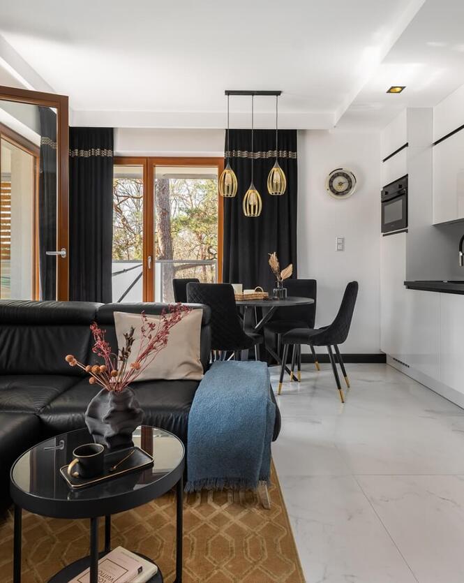 Nowoczesny apartament: biała kuchnia na wysoki połysk, czarna narożna sofa, złote lampy, jadalnia i balkon z drzewami.