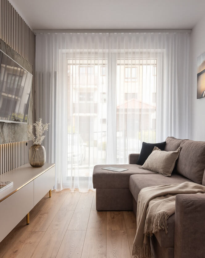 Przytulny apartamentowy salon z narożną kanapą, telewizorem, delikatnymi zasłonami, drewnianą podłogą i dekoracyjnym wazonem