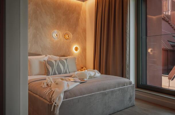Przytulny pokój hotelowy z tapicerowanym łóżkiem, wzorzystą poduszką, ciepłym nastrojowym oświetleniem, zasłonami i miejskim widokiem przez okno.