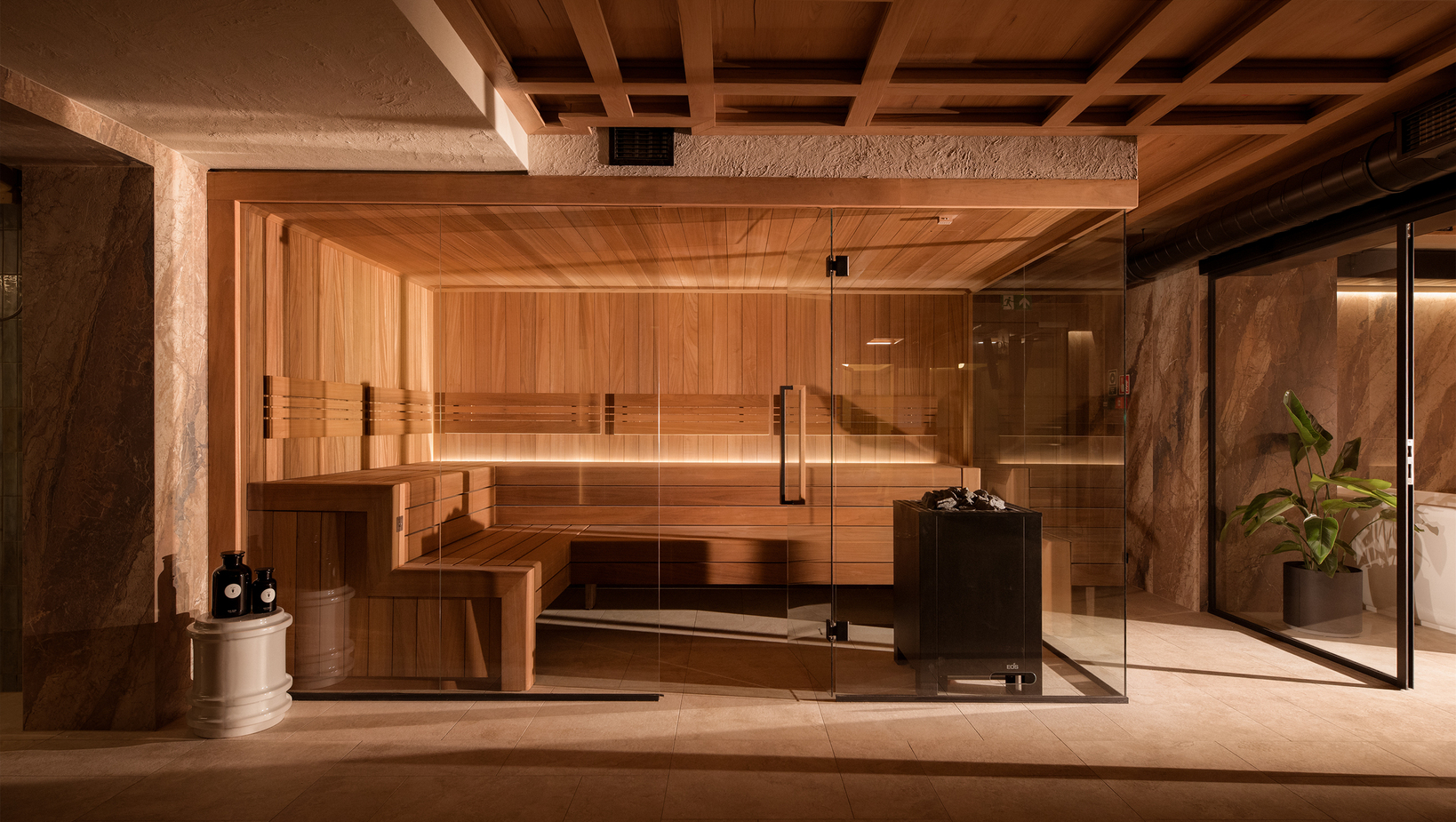 Sauna hotelowa z drewnianym wnętrzem, szklaną ścianą i podświetlanymi ławami; piec z kamieniami, kamienne ściany i roślina doniczkowa.