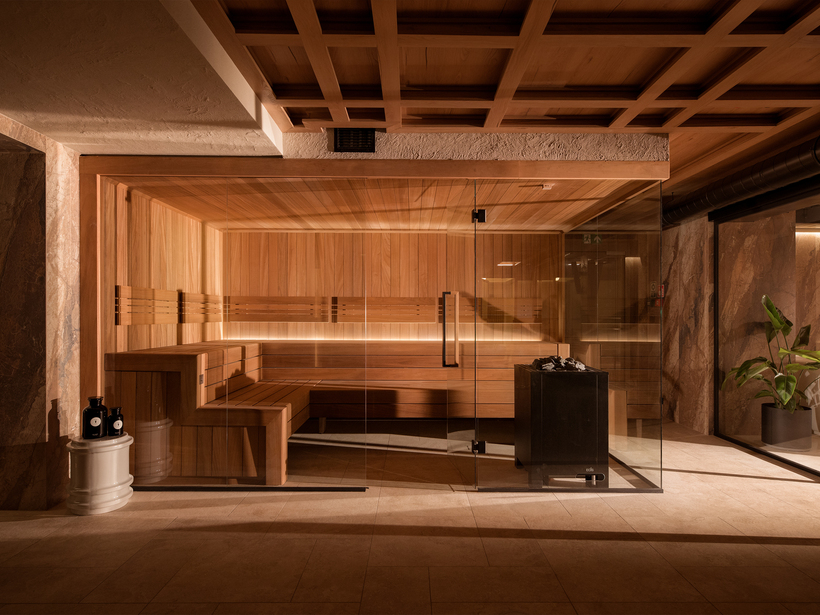 Sauna hotelowa z drewnianym wnętrzem, szklaną ścianą i podświetlanymi ławami; piec z kamieniami, kamienne ściany i roślina doniczkowa.