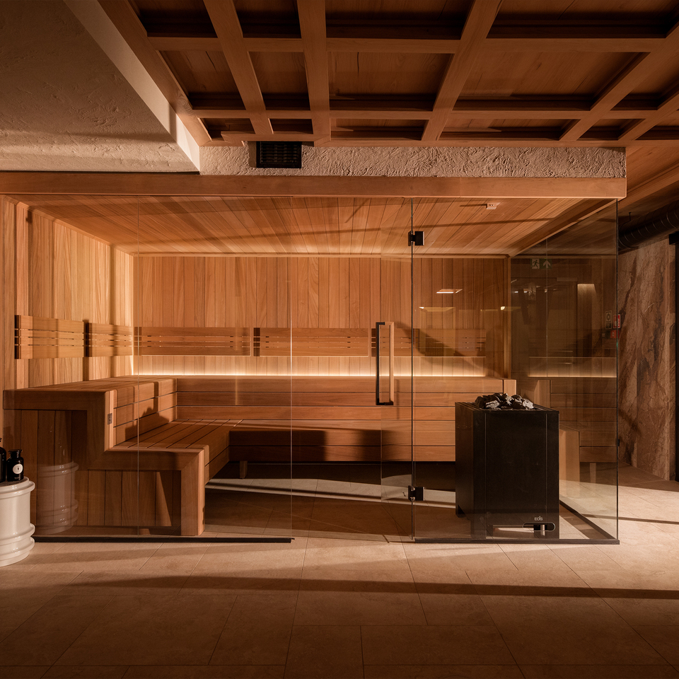 Sauna hotelowa z drewnianym wnętrzem, szklaną ścianą i podświetlanymi ławami; piec z kamieniami, kamienne ściany i roślina doniczkowa.