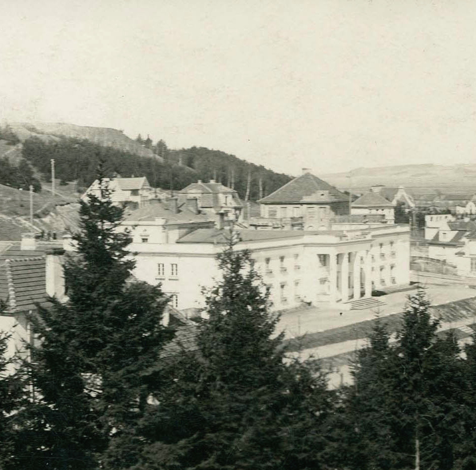 Klasyczny budynek z kolumnowym wejściem w górskiej miejscowości; wokół wille, iglaste drzewa i rozległa panorama wzgórz.