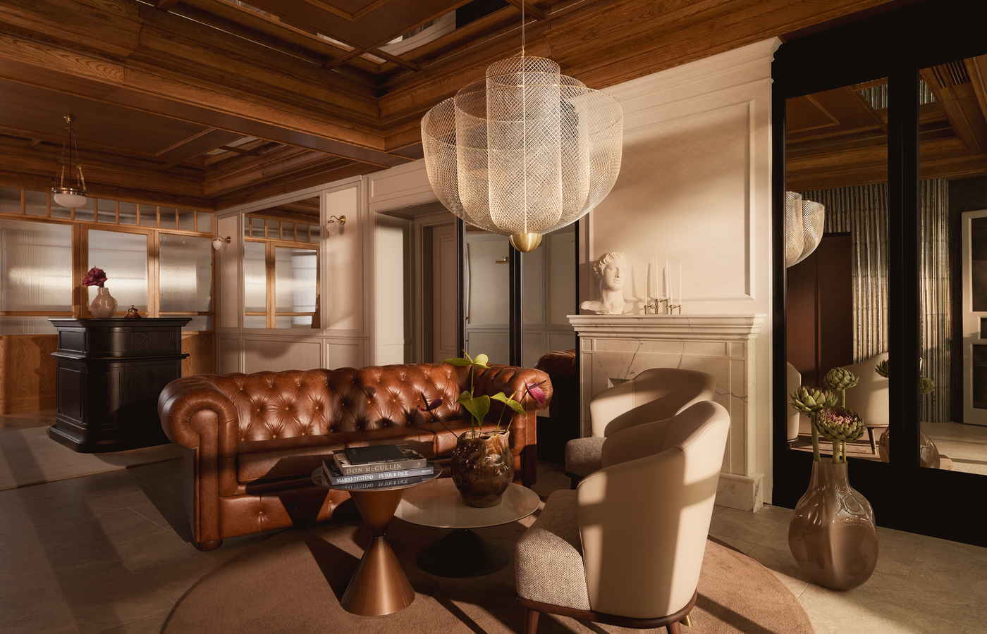 Przytulny salon hotelowy z pikowaną skórzaną sofą, dużym plecionym żyrandolem, marmurowym kominkiem z popiersiem i kasetonowym sufitem