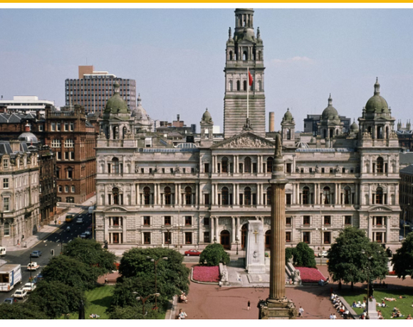 Grand neoclassical city hall facing a central column monument and green public square with lawns and surrounding historic buildings.