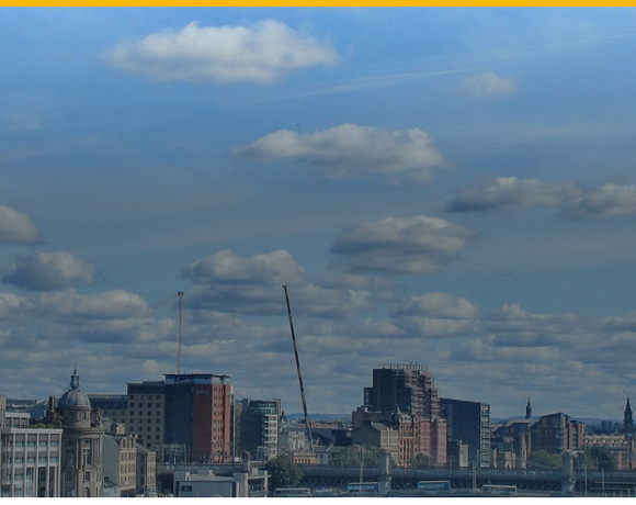 Panoramic city skyline with a mix of historic and modern buildings, a construction crane, and a bright blue sky dotted with fluffy clouds.