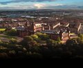 Aerial view of a grand red-sandstone landmark surrounded by parkland and trees, with river and city skyline in the distance, scenic urban vista