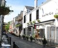 Cobbled lane with white pub, hanging flower baskets, string lights and outdoor café seating with potted plants.