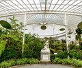 Glass-domed conservatory with ornate white ironwork, lush tropical plants and a white marble reclining statue on a pedestal