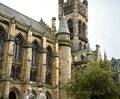 Gothic stone building with tall spire and arched windows, manicured lawn courtyard and two business travelers walking with briefcases