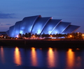 Curved modern waterfront venue lit blue at dusk with tree-lined promenade and orange streetlight reflections on the river.
