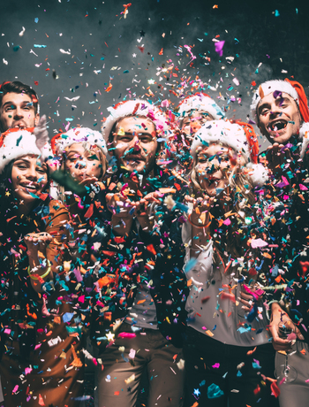 Hotel holiday party: smiling guests in Santa hats blowing colorful confetti, some holding drinks, lively festive atmosphere