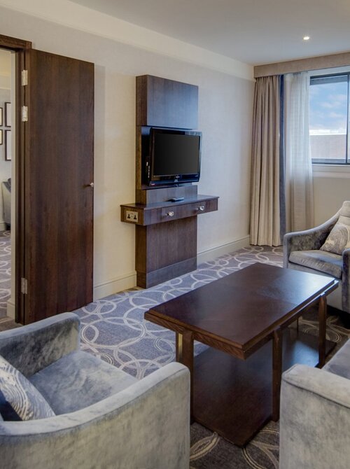 Spacious hotel suite with separate bedroom, king bed, plush seating area, TV, coffee table and city-view window.