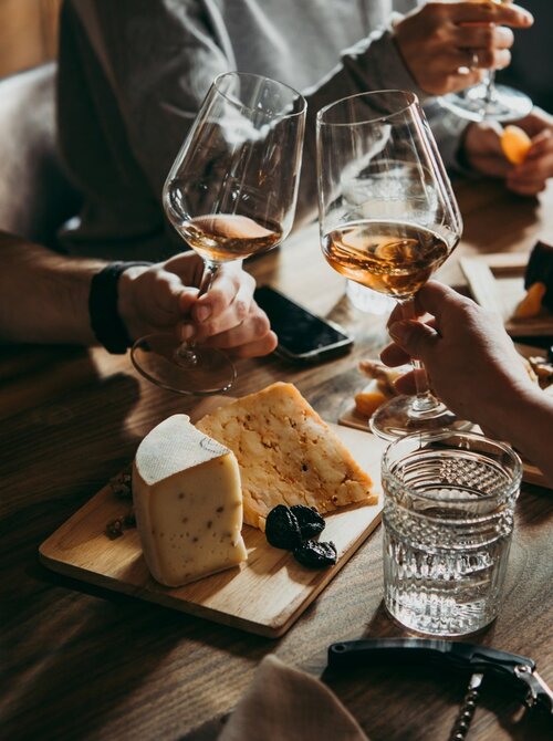 Friends toasting with amber wine over a rustic table, sharing an artisan cheese board with dried fruit, crystal water glasses and bottle.