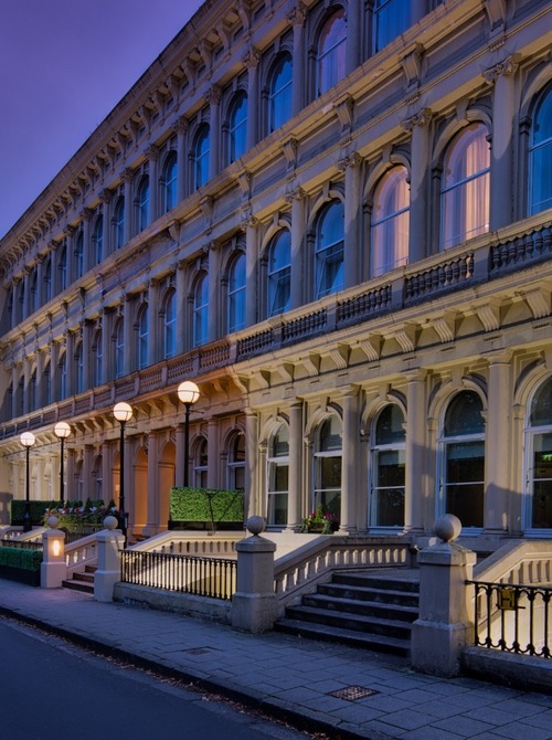 Historic hotel façade with arched windows, lit entrance steps and street lamps, city center beside church spire at dusk