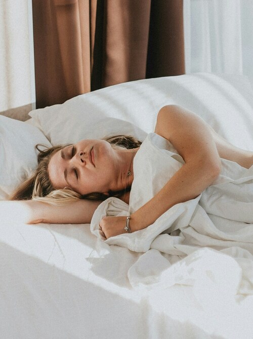 Woman sleeping in hotel bed as sunlight streams across crisp white linens, plush pillows and sheer curtains, relaxed morning.