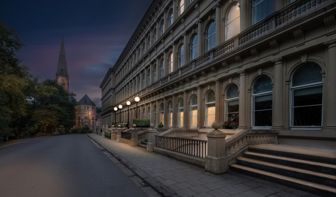 Grand hotel facade with arched windows, stone balustrades and steps lit by lamps at twilight, church spire beyond