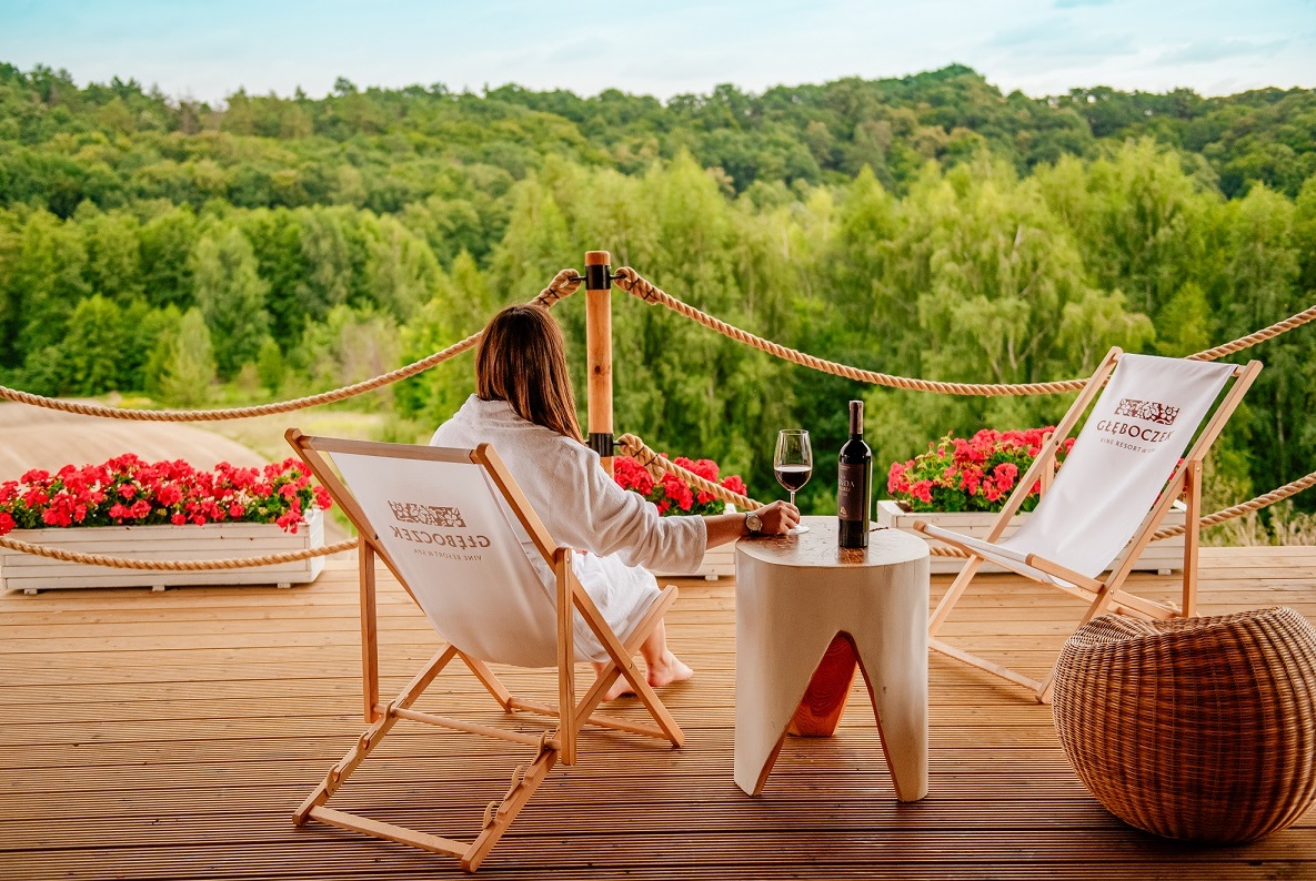 Gast im Bademantel auf Holzterrasse, genießt Rotwein am Tisch, mit Liegestühlen, Blumenkästen und Panoramablick auf den Wald