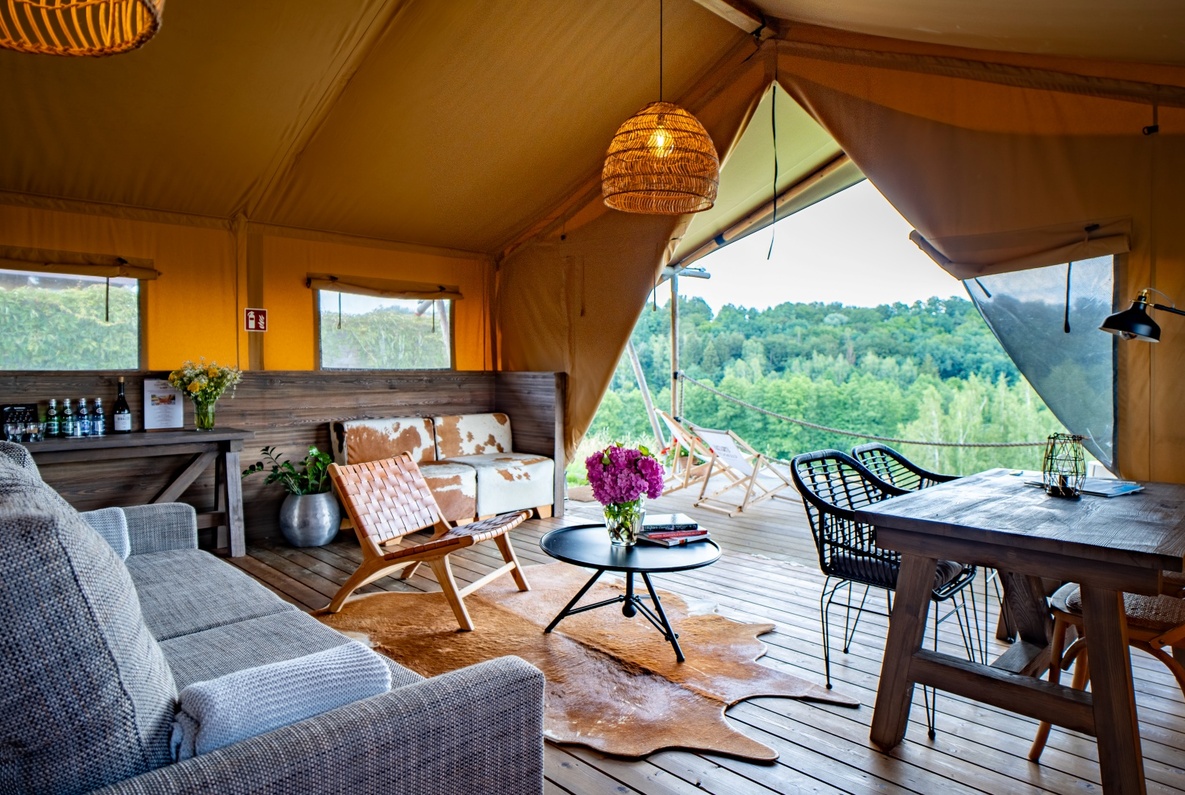 Spacious luxury glamping tent interior with comfortable furniture and a deck overlooking lush green forest views.