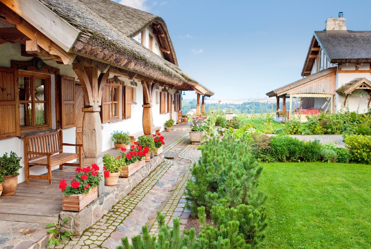 Reetgedecktes Landhotel mit Holzveranda, Sitzbank und roten Geranien in Kästen, Kopfsteinweg und gepflegtem Garten mit offener Feldsicht
