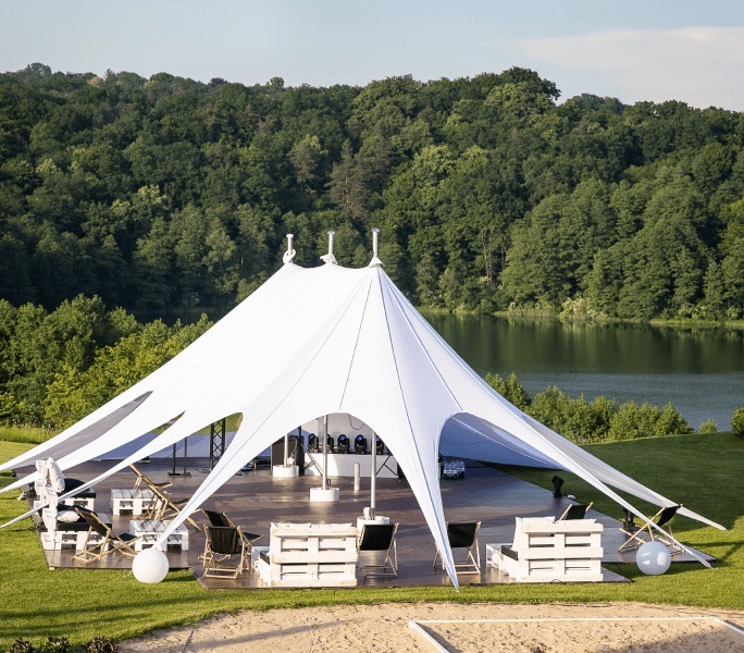 Spacious white event tent on a wooden deck by a lake, surrounded by green forest with comfortable wooden outdoor furniture.