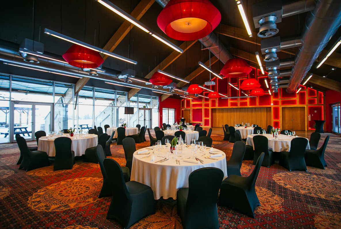 Spacious banquet hall with round tables dressed in white linens, black-covered chairs, red pendant lights, large windows and illuminated red feature wall