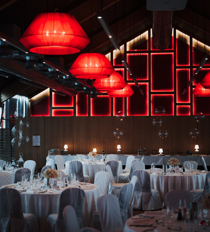 Banquet hall with round tables dressed in white linens and floral centerpieces, dramatic red pendant lamps and geometric red wall lighting