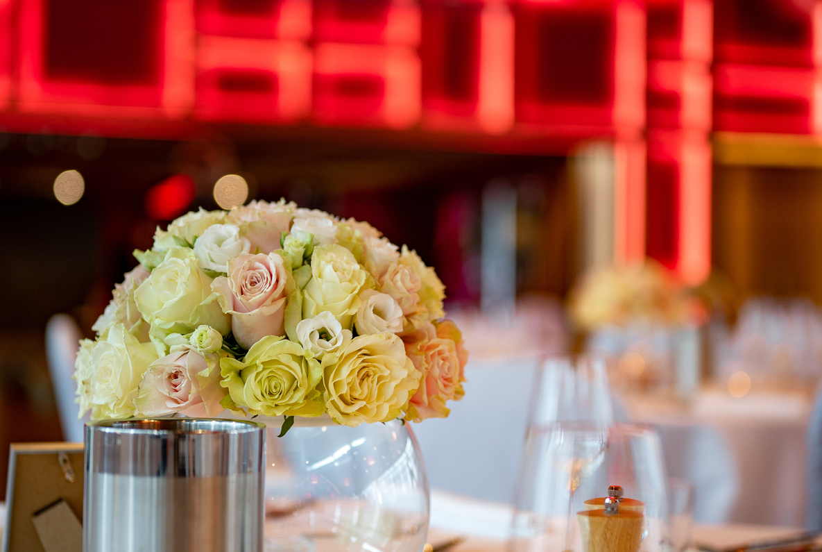 Glass-vase bouquet of pale roses on a white table with wine cooler, pepper mill and elegant place settings, red neon backdrop