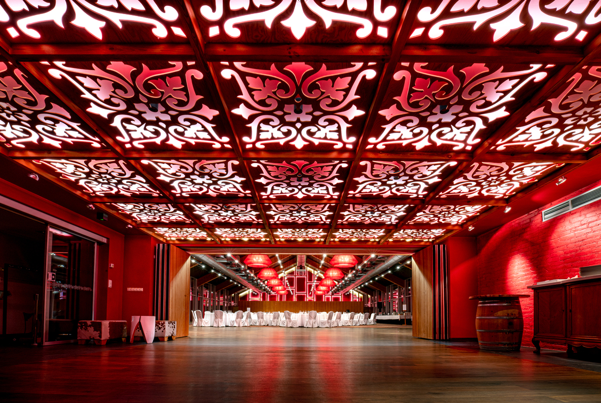 Ornate backlit ceiling panels and red-lit banquet hall with rows of white-clothed tables, wooden floor and barrel decor.