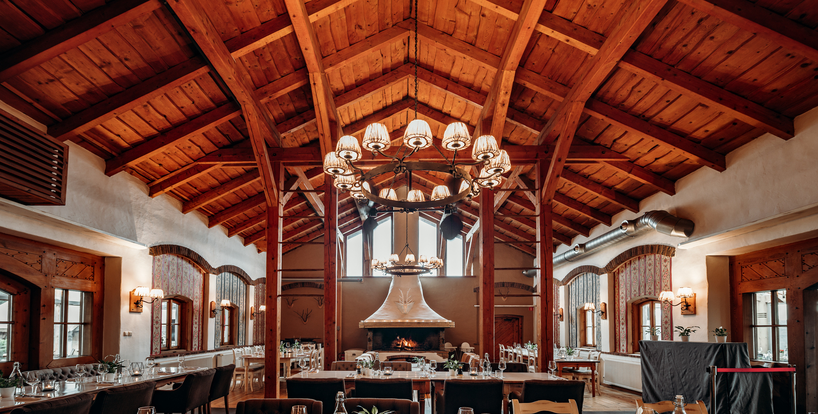 Rustic dining hall with vaulted wooden beams, central chandelier and large fireplace; tables set for hotel dinner.