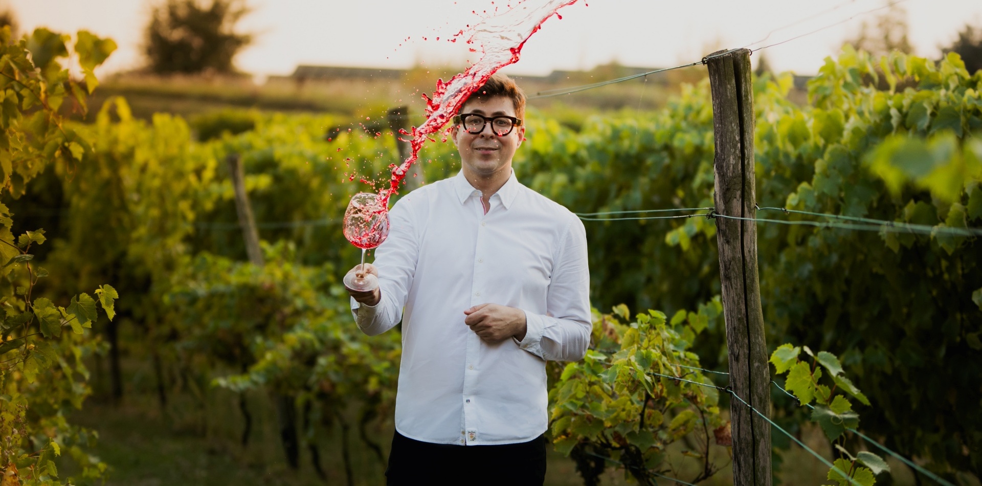 Mężczyzna w białej koszuli rzuca kieliszkiem czerwonego wina w winnicy podczas złotej godziny.