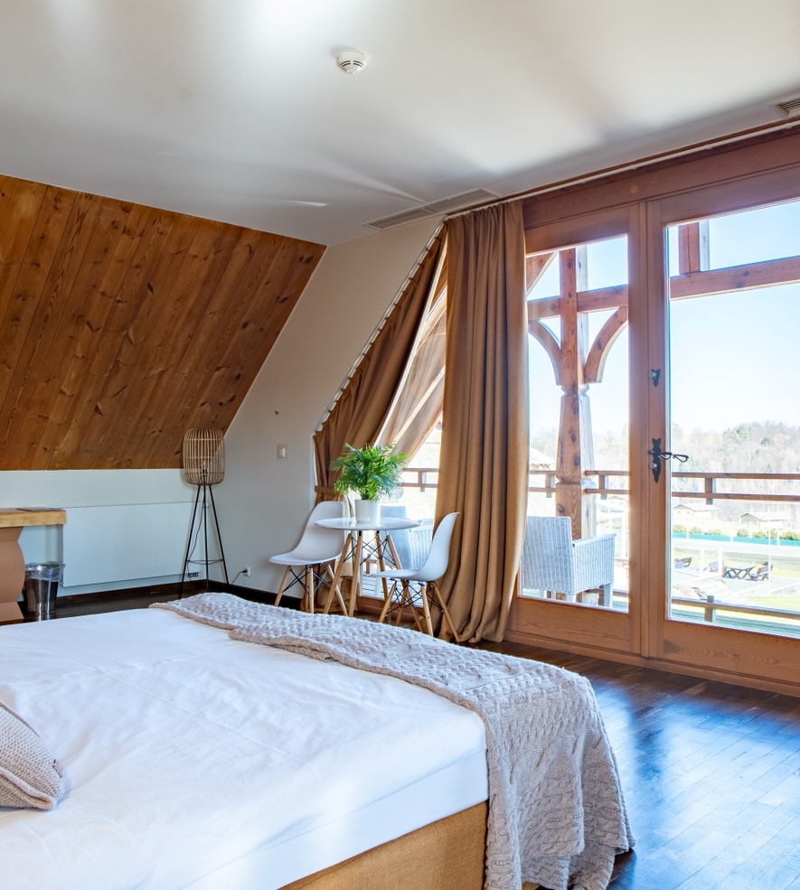 Przytulny pokój hotelowy na poddaszu z dużym łóżkiem, drewnianą ścianą i balkonem z widokiem na zielony krajobraz.