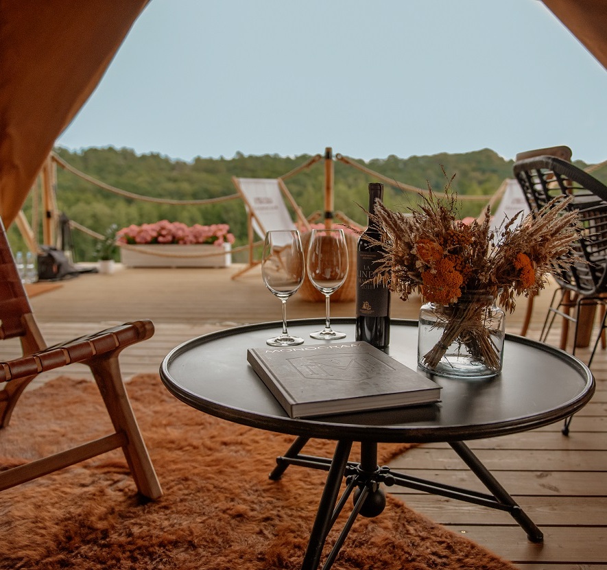Cozy wooden gazebo with comfortable chairs, table set with wine and flowers, overlooking lush green hills.