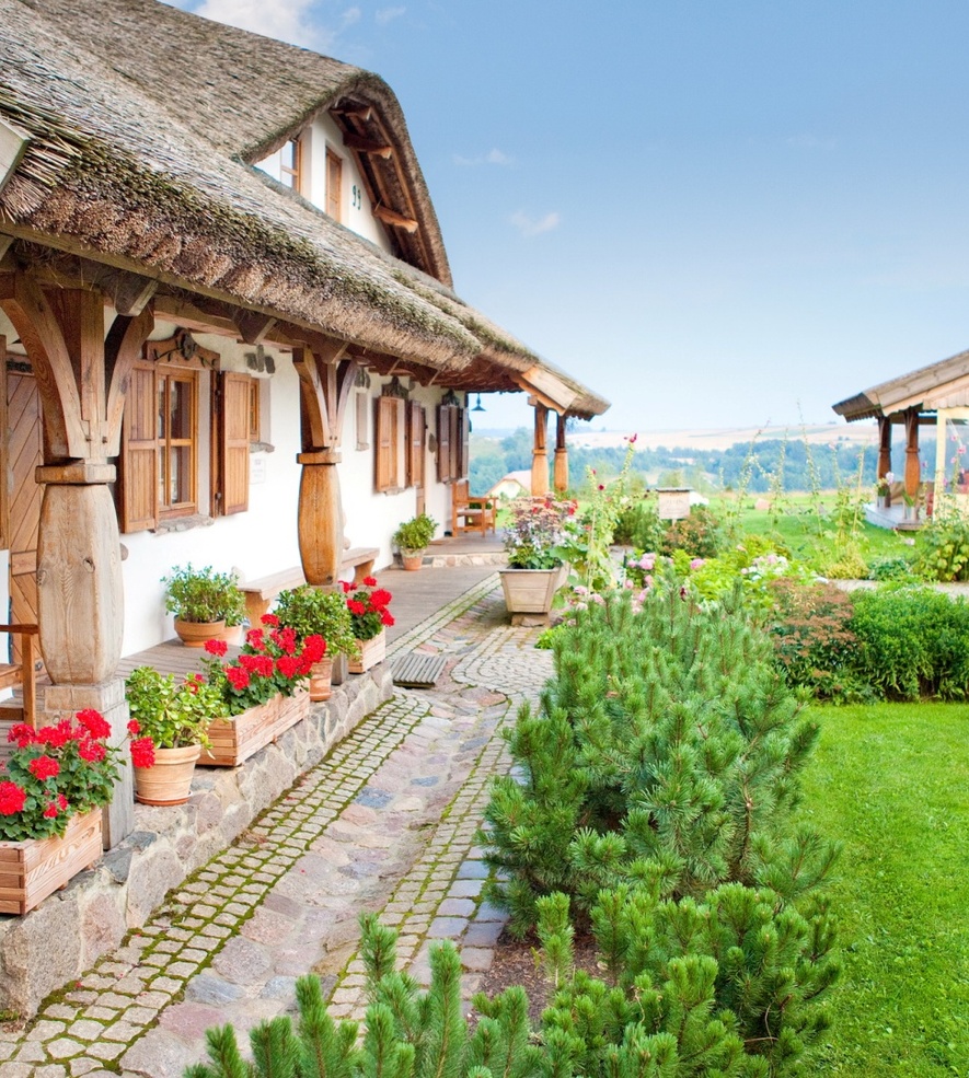 Rustic hotel with thatched roof porch, wooden benches, potted flowers, and a lush green garden in a countryside setting