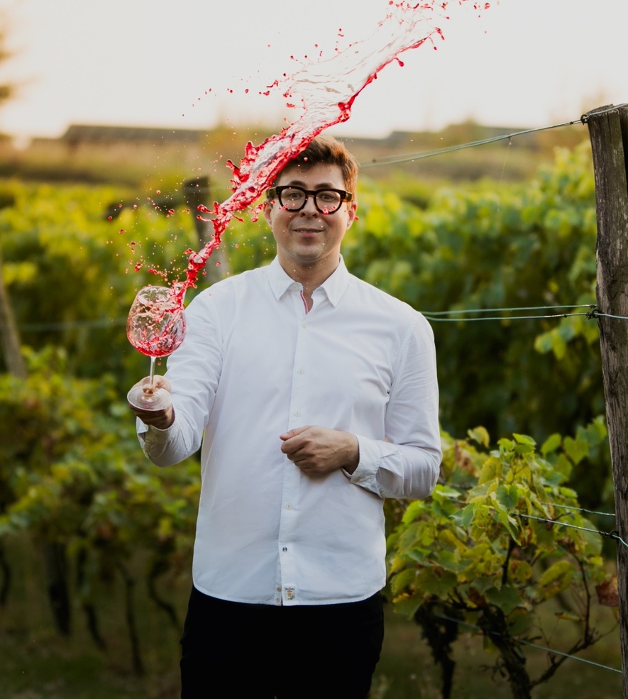 Man in a white shirt tossing a glass of red wine in a vineyard at golden hour.