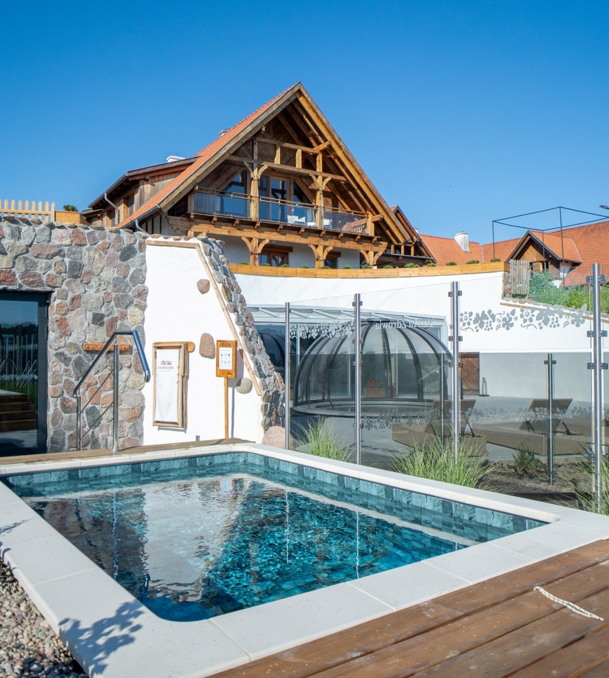 Outdoor heated pool beside a stone wall and traditional wooden building with balcony under clear blue sky.
