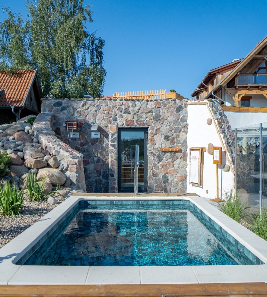 Outdoor stone pool by hotel spa with sauna, surrounded by greenery and wooden building in the background